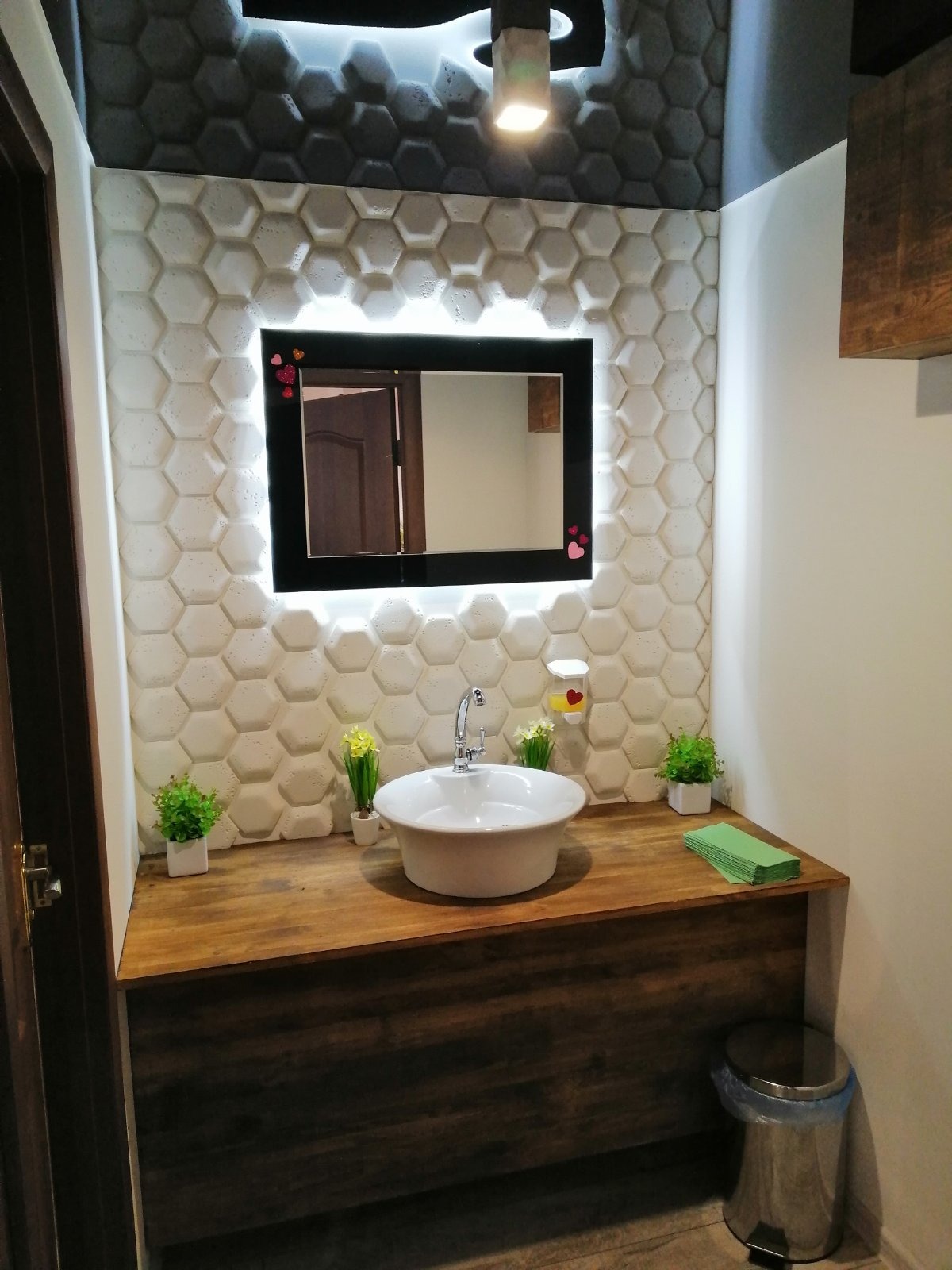 Nowoczesna łazienka z umywalką nablatową na drewnianym blacie, heksagonalne płytki ścienne z podświetleniem LED wokół lustra, wiszący dozownik do mydła z symbolem serca, dekoracyjne rośliny...