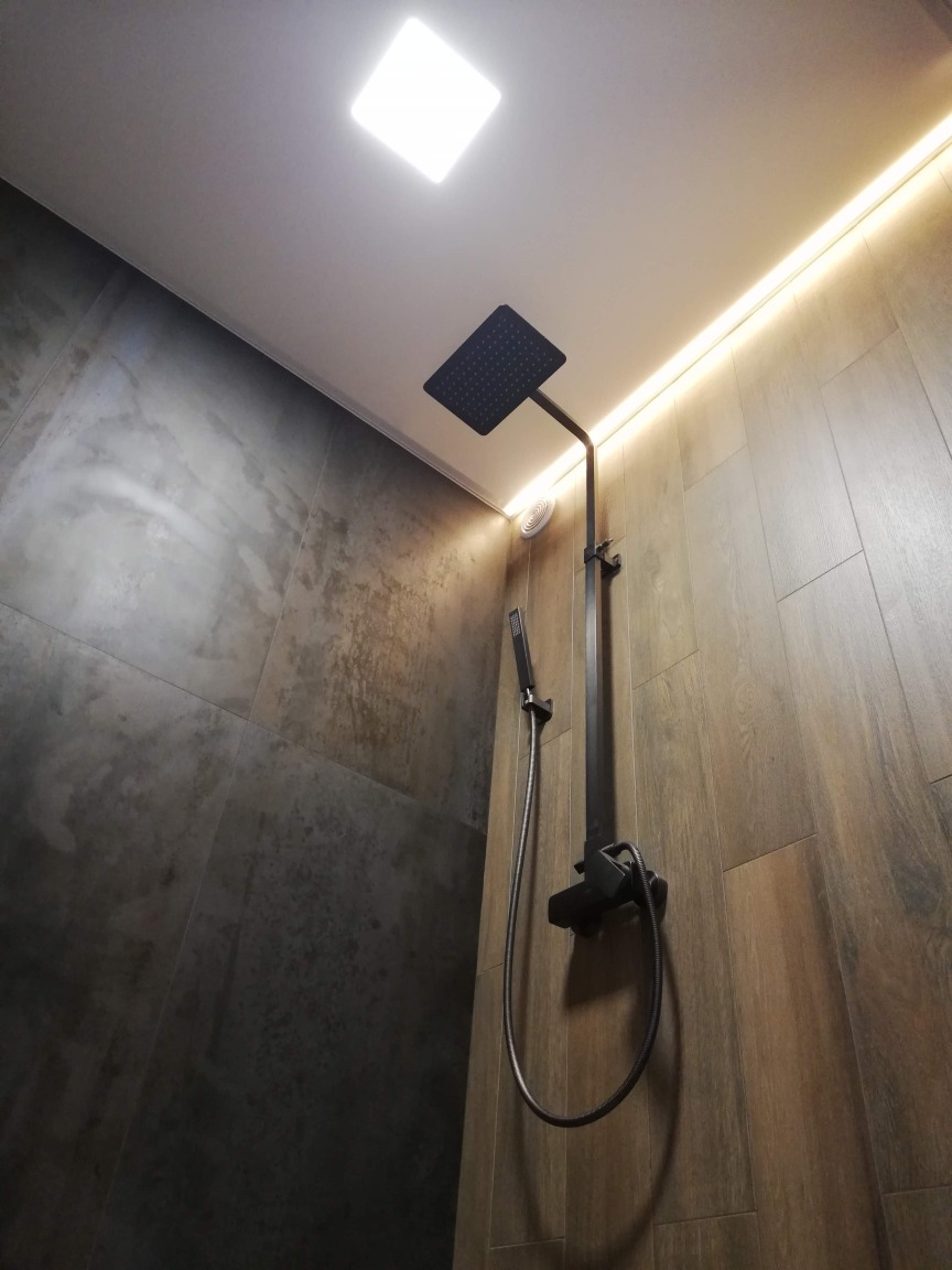 Nowoczesna łazienka z ciemnymi, drewnopodobnymi płytkami, czarną armaturą prysznicową i oświetleniem LED pod sufitem.