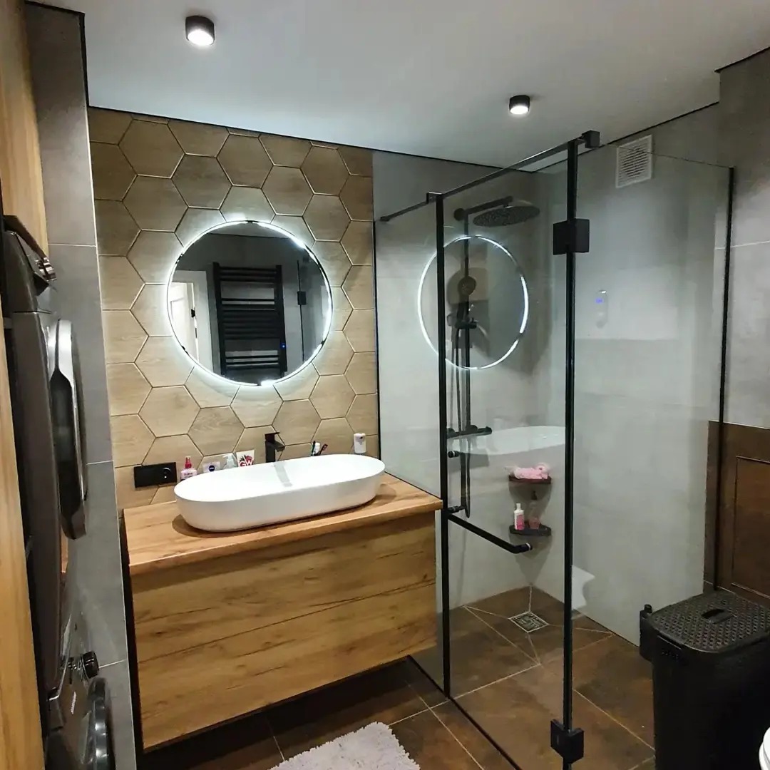 Nowoczesna łazienka z heksagonalnymi płytkami, okrągłym lustrem z podświetleniem LED, umywalką nablatową na drewnianej szafce i kabiną prysznicową typu walk-in z czarnymi profilami.