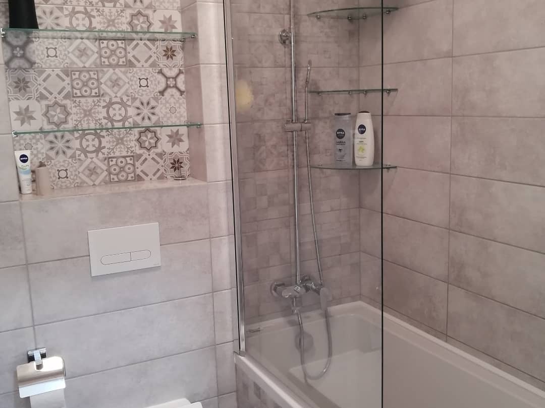 Jasna łazienka z wanną za szklanym parawanem, ozdobnymi płytkami w niszy z półkami, oraz minimalistyczną armaturą.