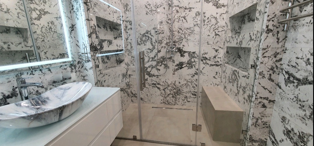 Nowoczesna łazienka z marmurowymi płytkami, szklaną kabiną prysznicową i umywalką nablatową o wzorze marmuru, oświetlone lustro i wnęki w ścianie.