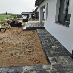 TOM-BRUK Tomasz Rogoziński usługi ogólnobudowlane - Częściowo ułożona ścieżka z szaro-czarnej kostki brukowej prowadząca do domu, z dwoma pracownikami odpoczywającymi na krzesłach w tle, obok sterty kostki i zabawek pluszowych.