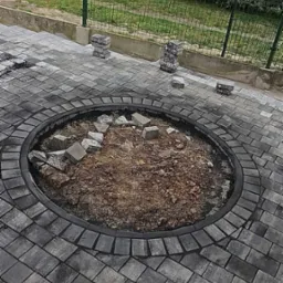 Okragłe miejsce na ognisko lub rabatę kwiatową w trakcie budowy, otoczone szarą kostką brukową, z widocznym fragmentem zagęszczarki w rogu oraz zielonym ogrodzeniem panelowym w tle.