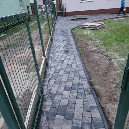 TOM-BRUK Tomasz Rogoziński usługi ogólnobudowlane - Ścieżka z kostki brukowej w Wałczu, prowadząca od furtki ogrodzenia do budynku, z paletą na trawniku i przechodzącą osobą w tle.