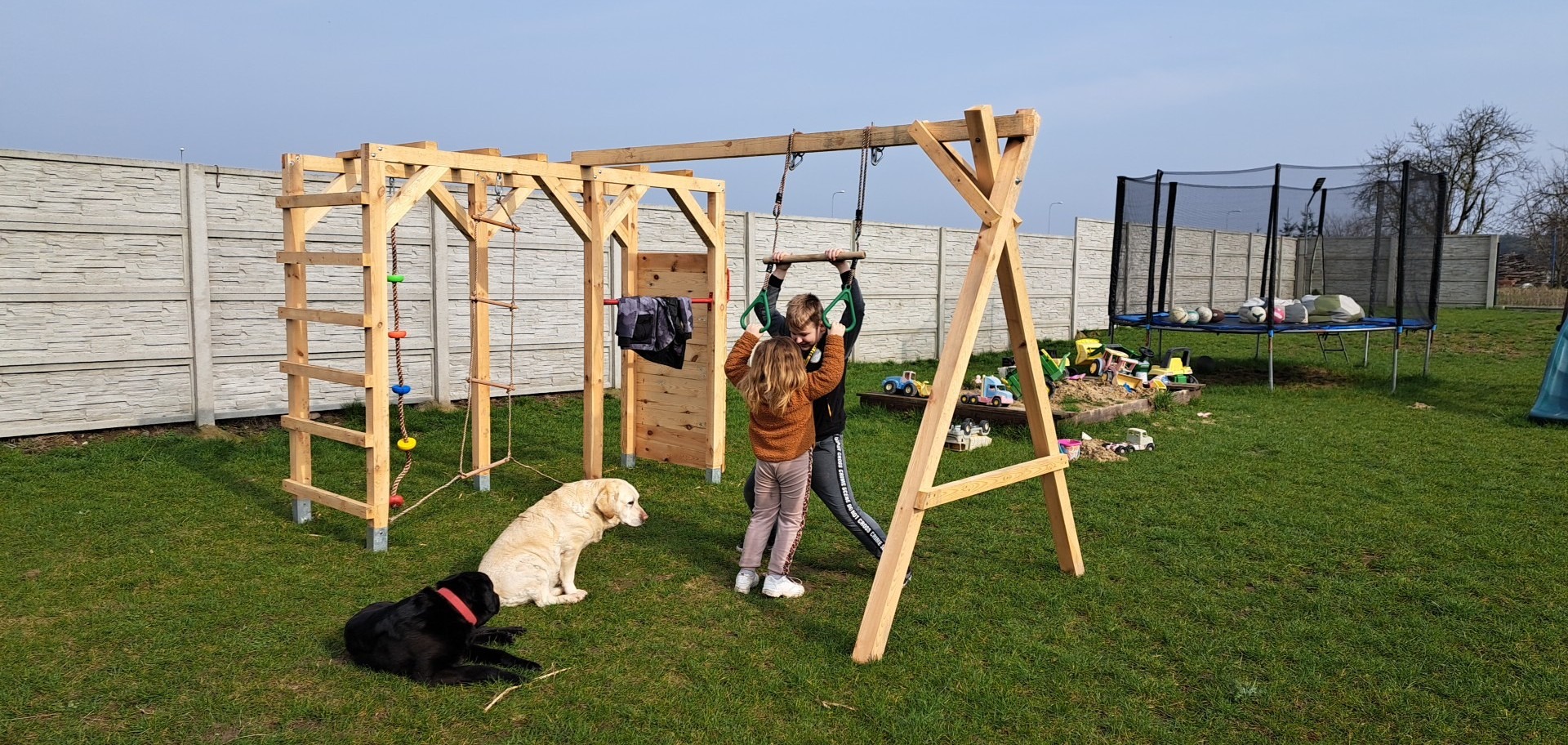 Dzieci bawią się na drewnianym placu zabaw z drabinkami, linami i huśtawką na tle betonowego ogrodzenia, trampolina w tle, dwa psy w pobliżu.