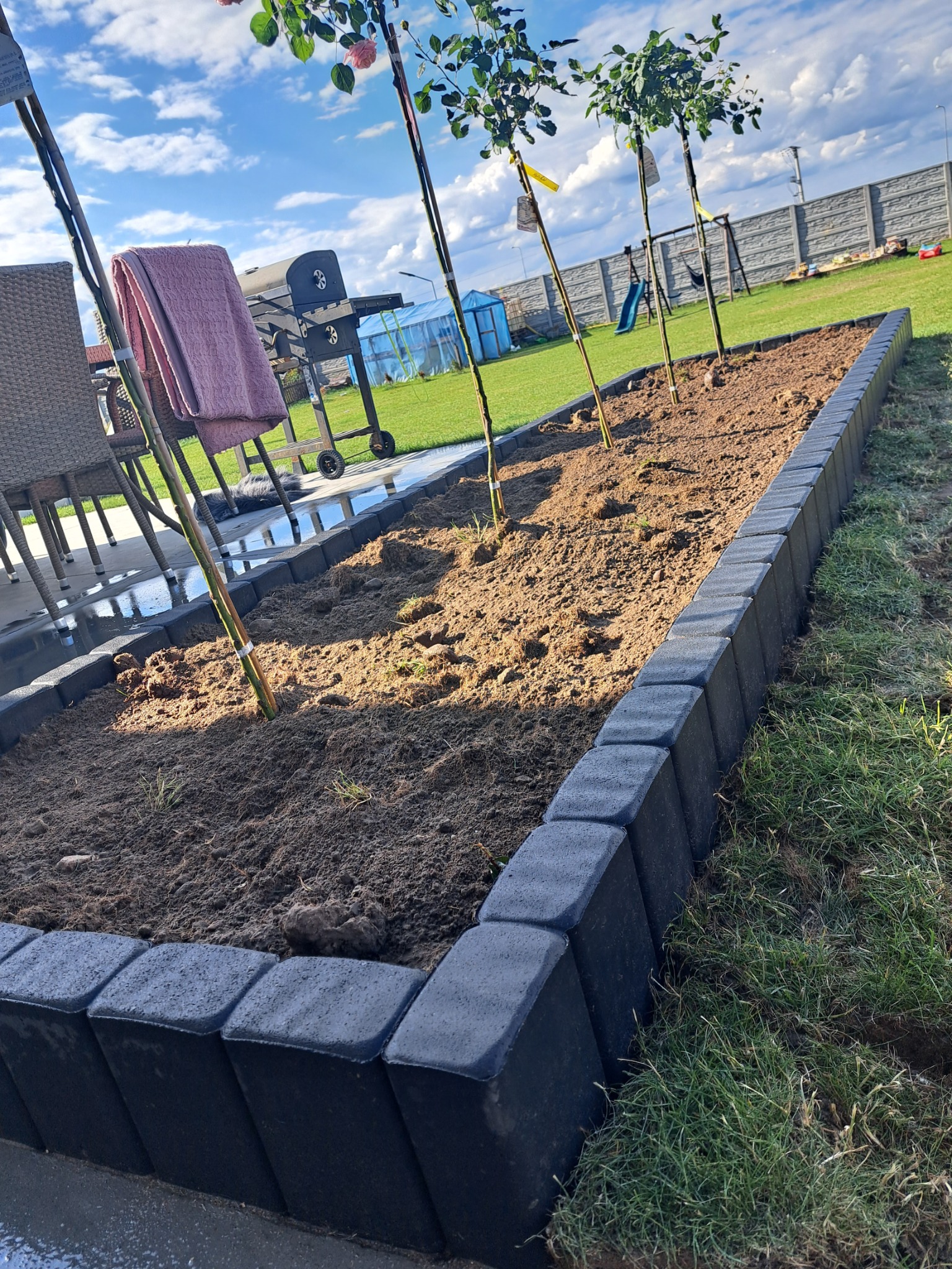 Krawężnik z betonowych kostek obramowujący rabatę z posadzoną młodą roślinnością, widok na ogród z grillem i placem zabaw w tle.