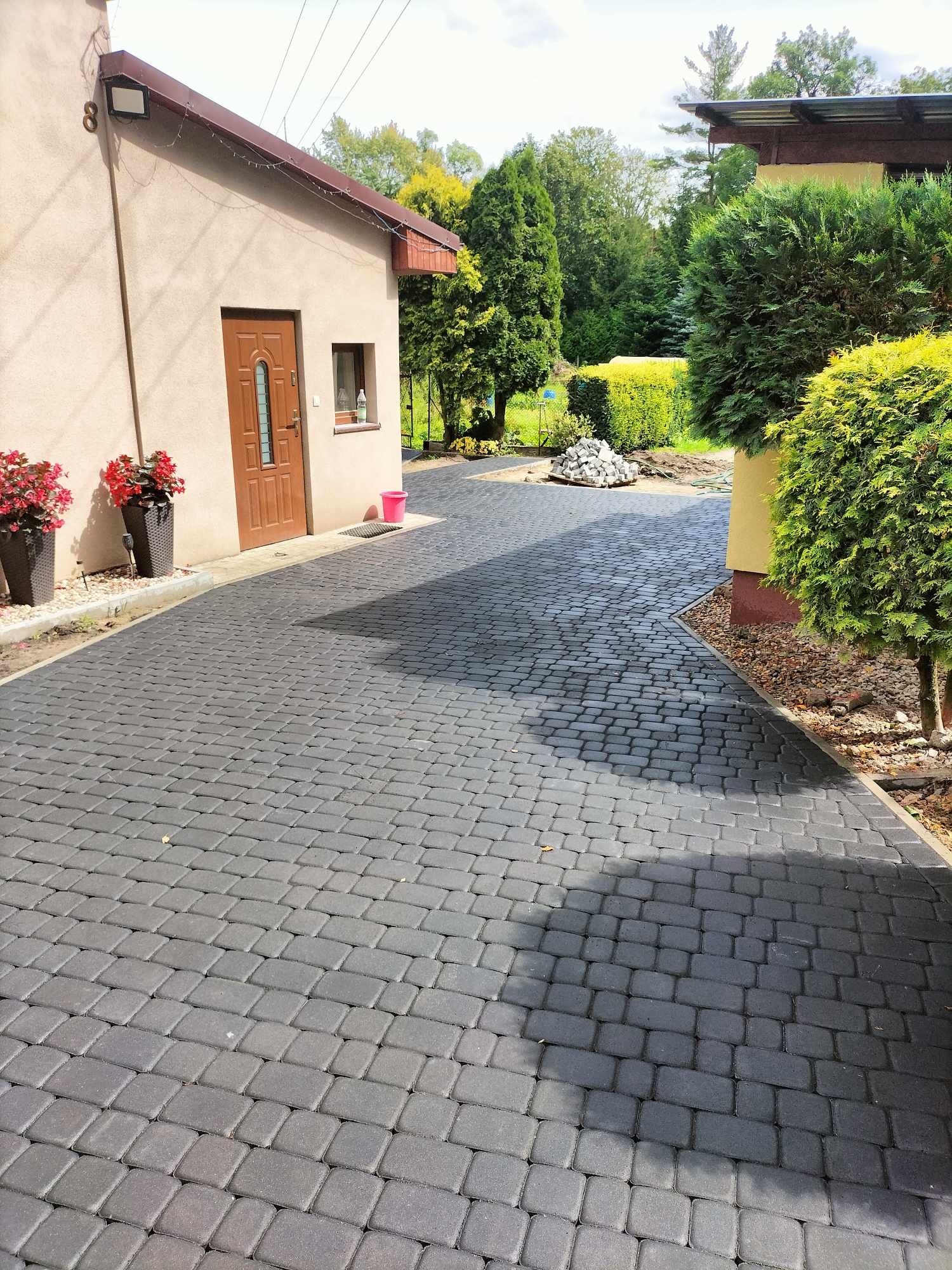 Ułożona, ciemnoszara kostka brukowa prowadząca do domu, z widocznymi donicami z czerwonymi kwiatami przy wejściu i stertą kostki brukowej w oddali.  Widoczny fragment ogrodu z krzewami.