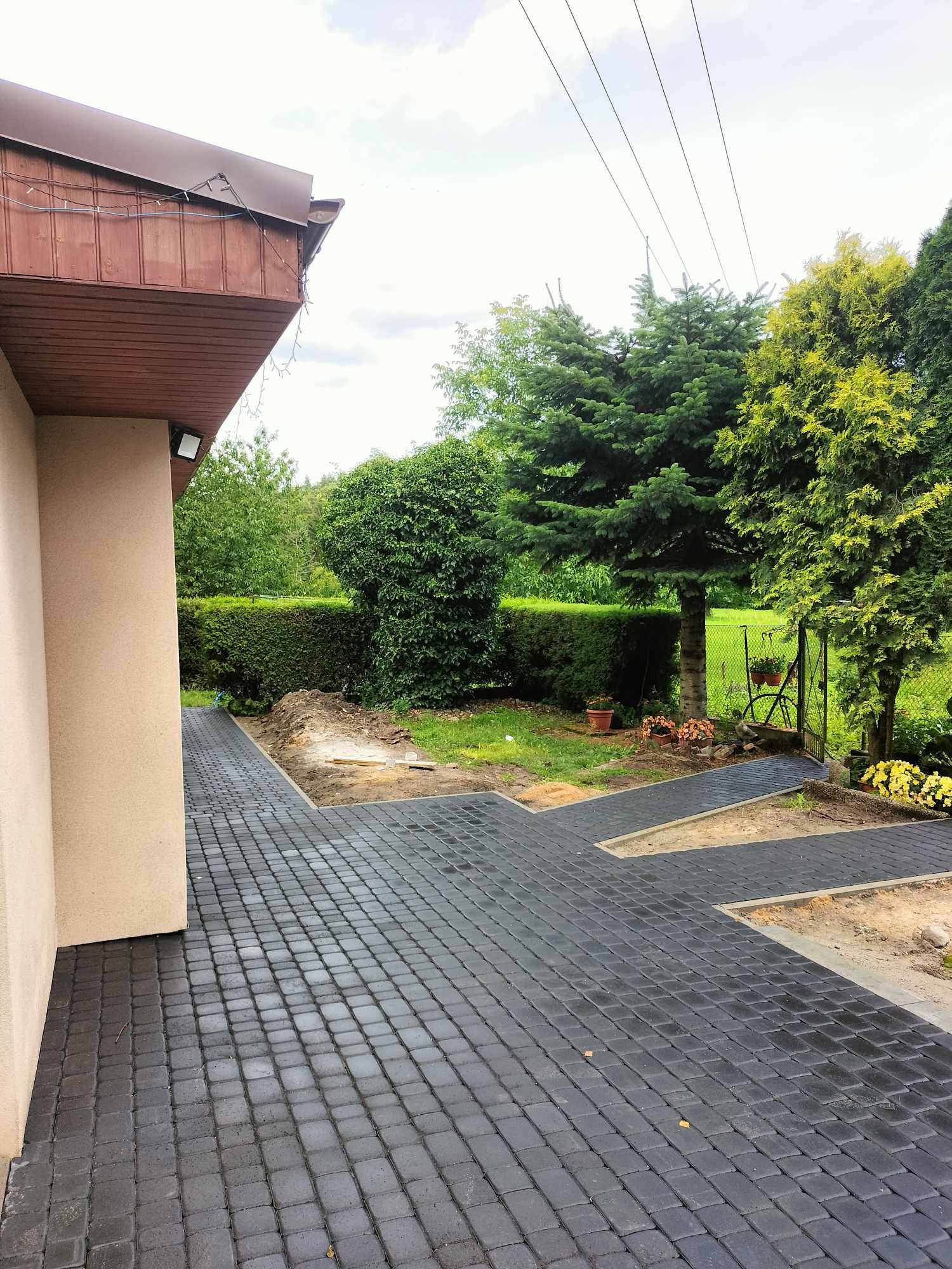 Ułożona, grafitowa kostka brukowa tworząca ścieżkę w ogrodzie, prowadząca od ściany domu do furtki, z widocznymi rabatami kwiatowymi po bokach i zielenią w tle.