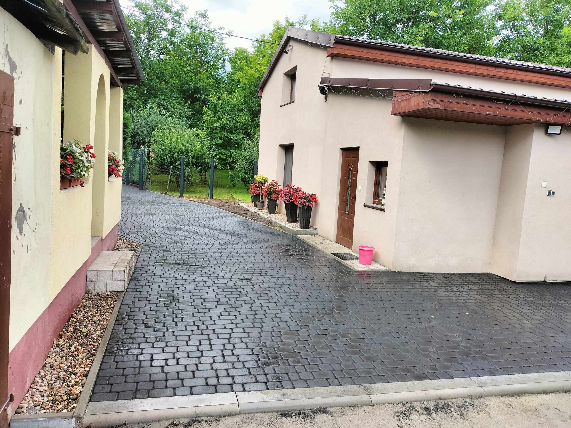 Ułożona, ciemnoszara kostka brukowa prowadząca do domu z beżową elewacją, kwiaty w donicach i różowe wiaderko przy wejściu.