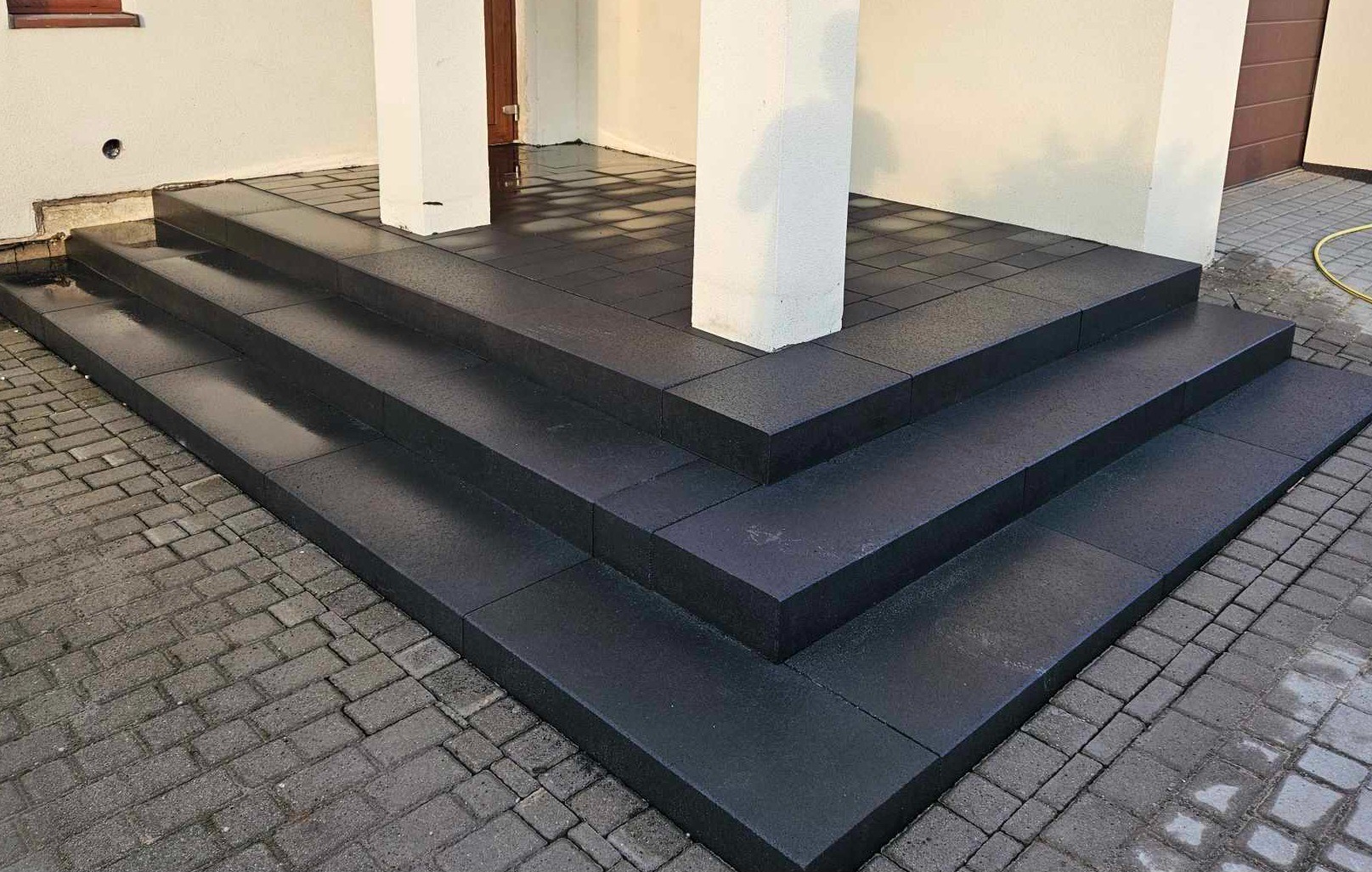 Schody wejściowe wyłożone ciemną kostką brukową, prowadzące do budynku z białymi kolumnami, po deszczu, z widoczną kostką brukową na podejściu.