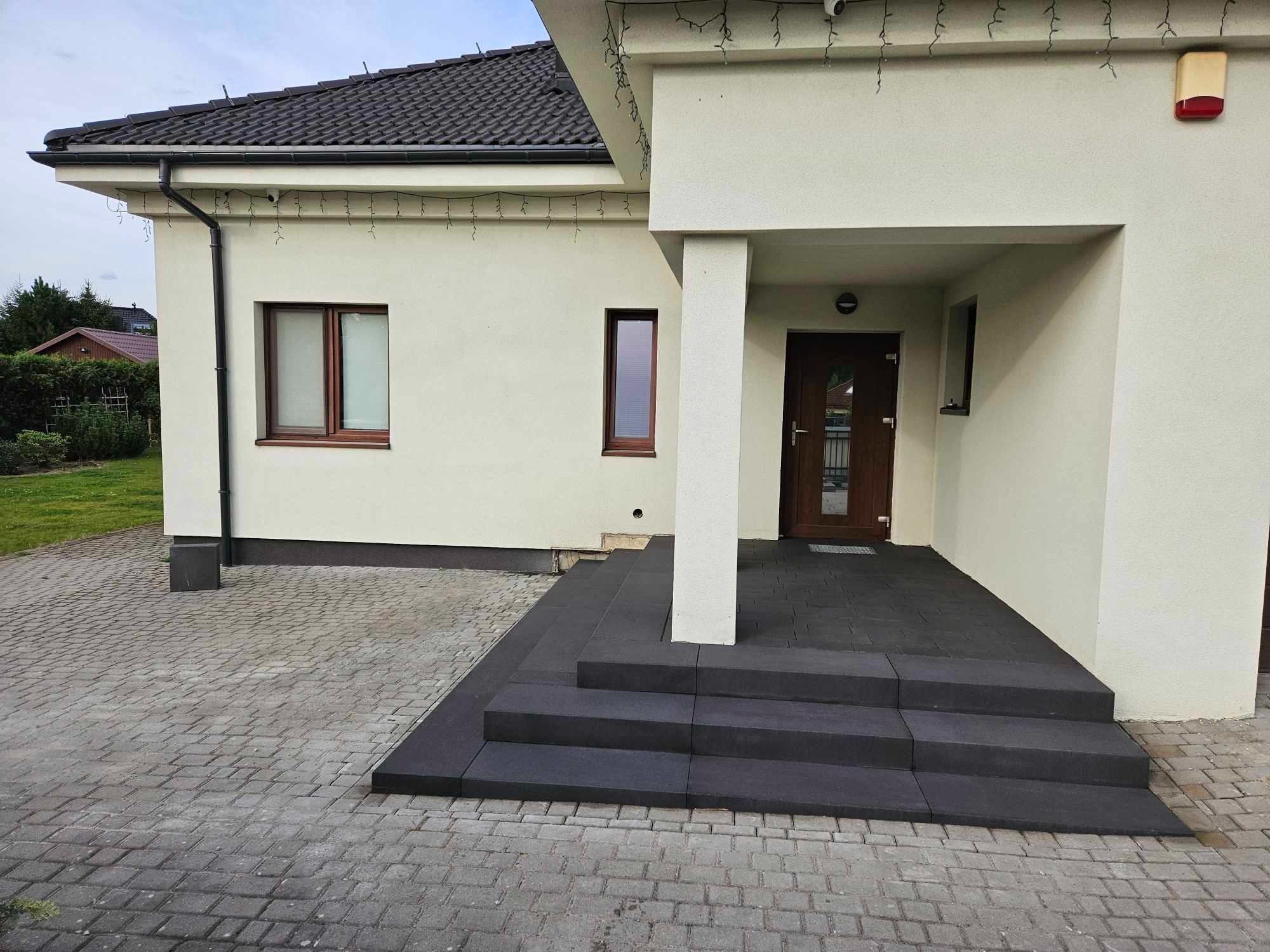 Nowoczesne schody wejściowe z ciemnej kostki brukowej prowadzące do domu z jasną elewacją i brązowymi oknami, otoczone kostką brukową w odcieniach szarości.