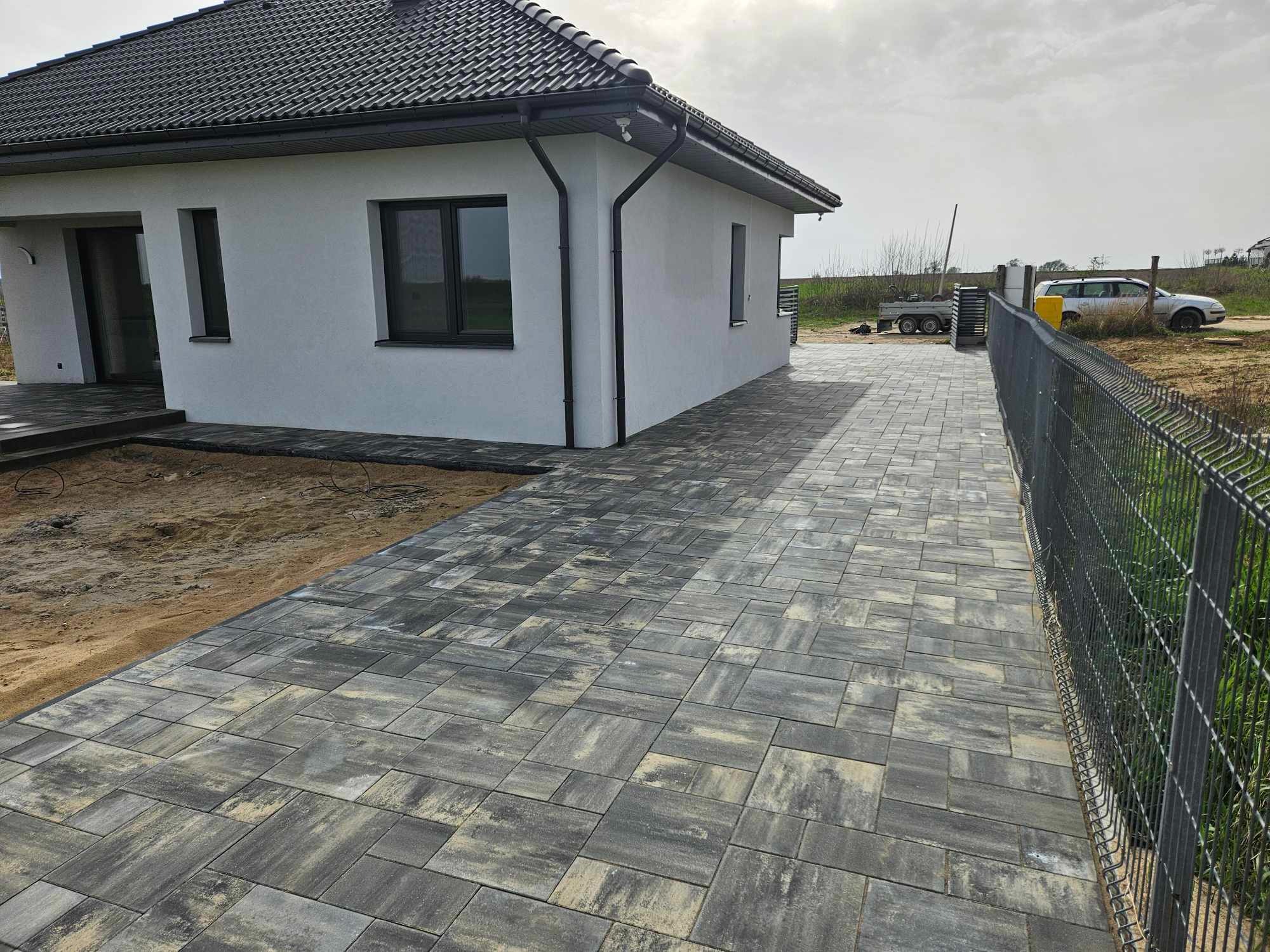 Ułożona szara kostka brukowa prowadząca od domu z ciemną stolarką okienną i dachem do metalowego ogrodzenia siatkowego, widoczny fragment tarasu i trawnika w tle.