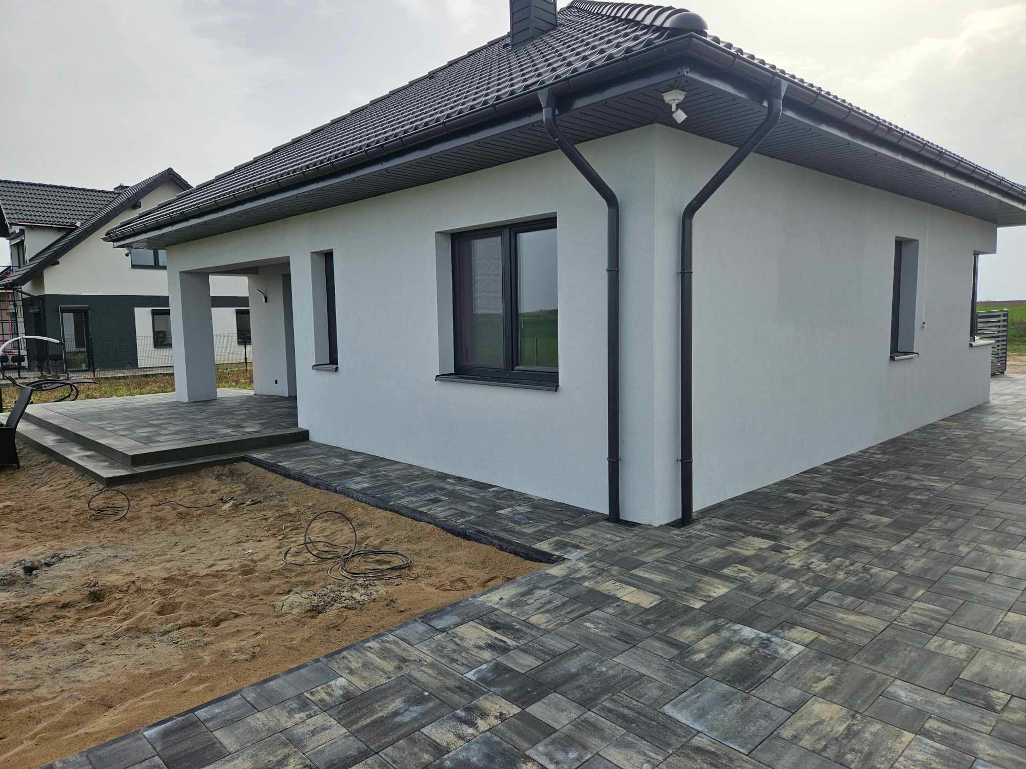 Nowoczesny dom jednorodzinny z ciemną dachówką i szarą kostką brukową na podjeździe i ścieżce, widoczne przewody elektryczne w piasku przed domem.