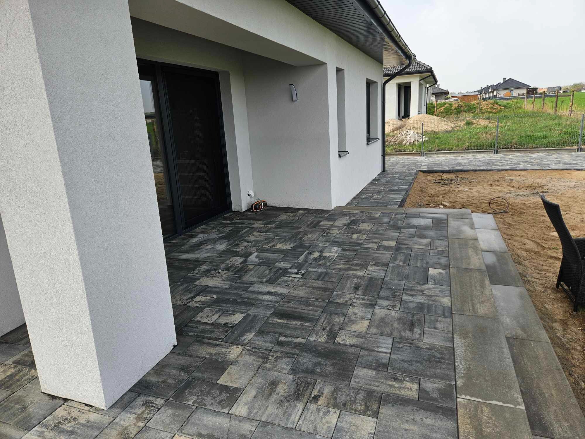 Nowoczesny taras z szarej kostki brukowej ułożonej wzorem jodełki, przylegający do białego domu z dużymi oknami, widok na ogród w tle.