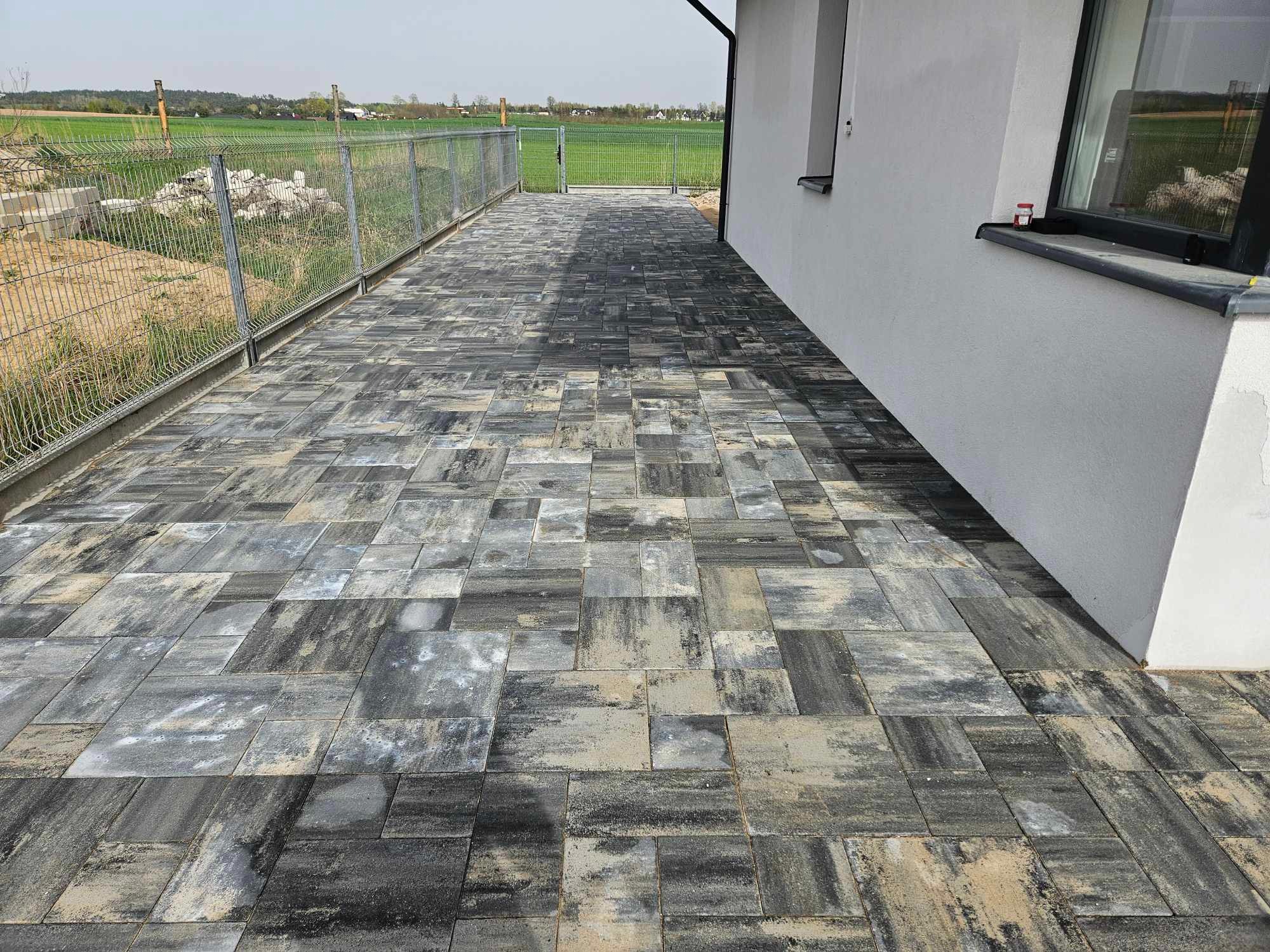Nowo ułożony chodnik z kostki brukowej o nieregularnym wzorze i szaro-beżowej kolorystyce, przylegający do białego budynku z widocznym oknem i ogrodzeniem z siatki w tle.