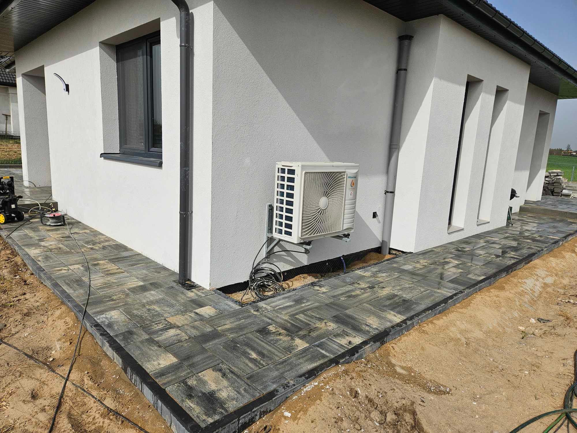 Ścieżka z kostki brukowej w odcieniach szarości, obramowana czarnym obrzeżem, prowadząca wzdłuż białej ściany domu z klimatyzatorem i szarą rynną, widoczny piasek i narzędzia.