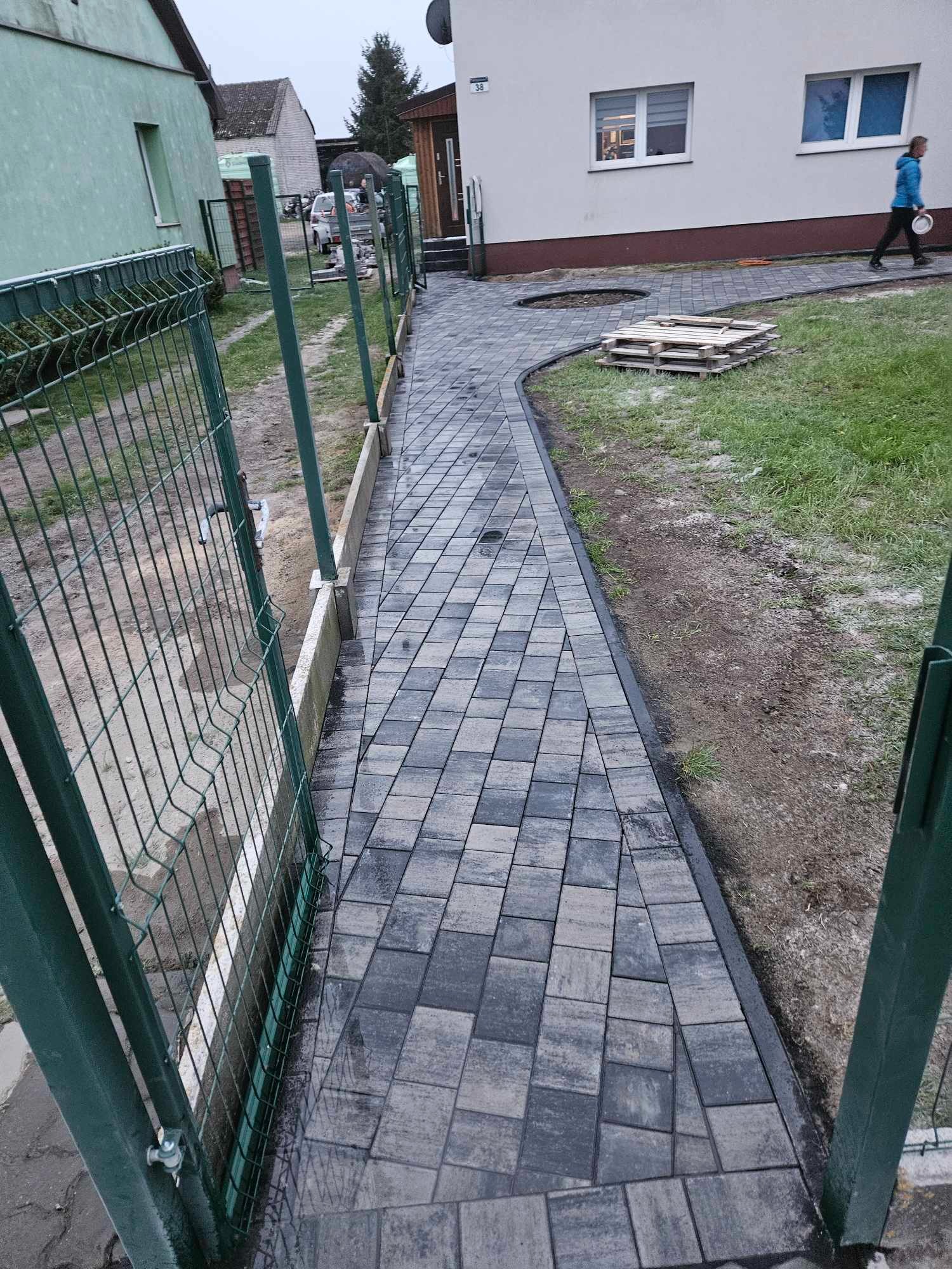 Ścieżka z kostki brukowej w Wałczu, prowadząca od furtki ogrodzenia do budynku, z paletą na trawniku i przechodzącą osobą w tle.