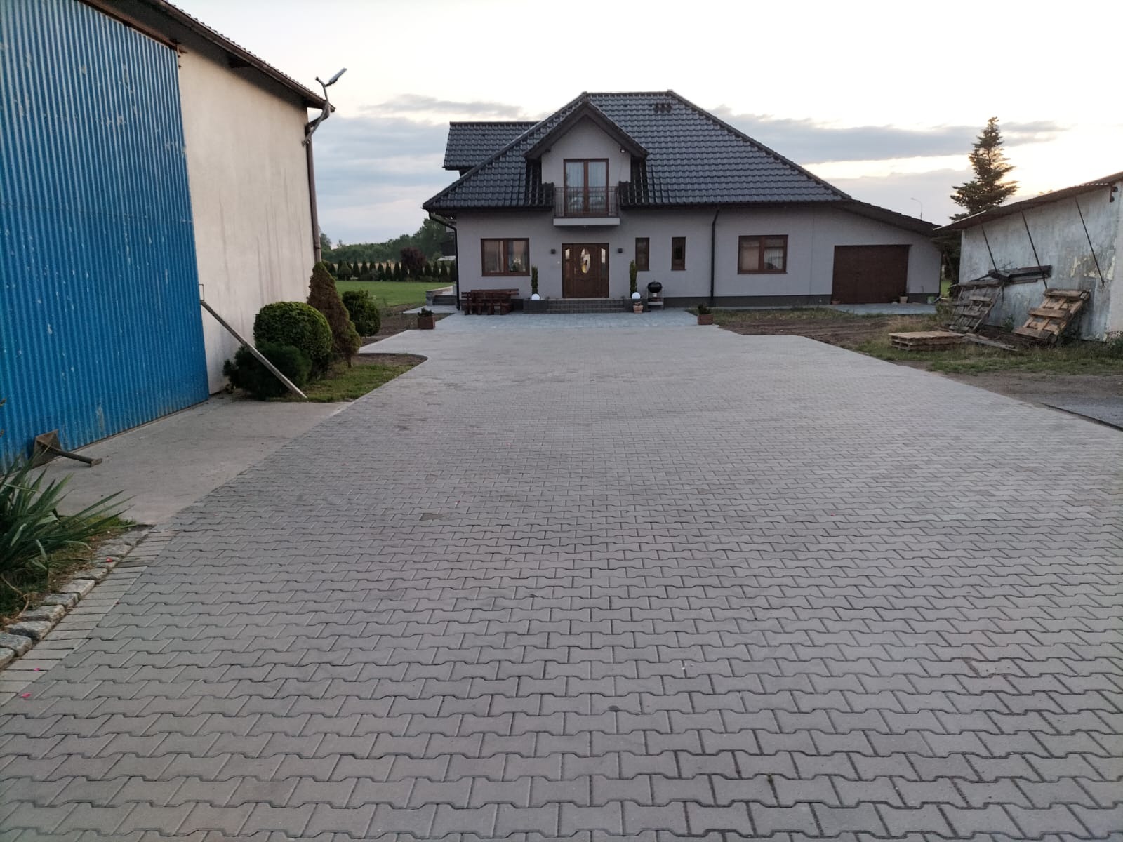 Szeroki podjazd z szarej kostki brukowej w kształcie litery I, prowadzący do nowoczesnego domu z szarą elewacją i ciemnym dachem, widok z poziomu gruntu.