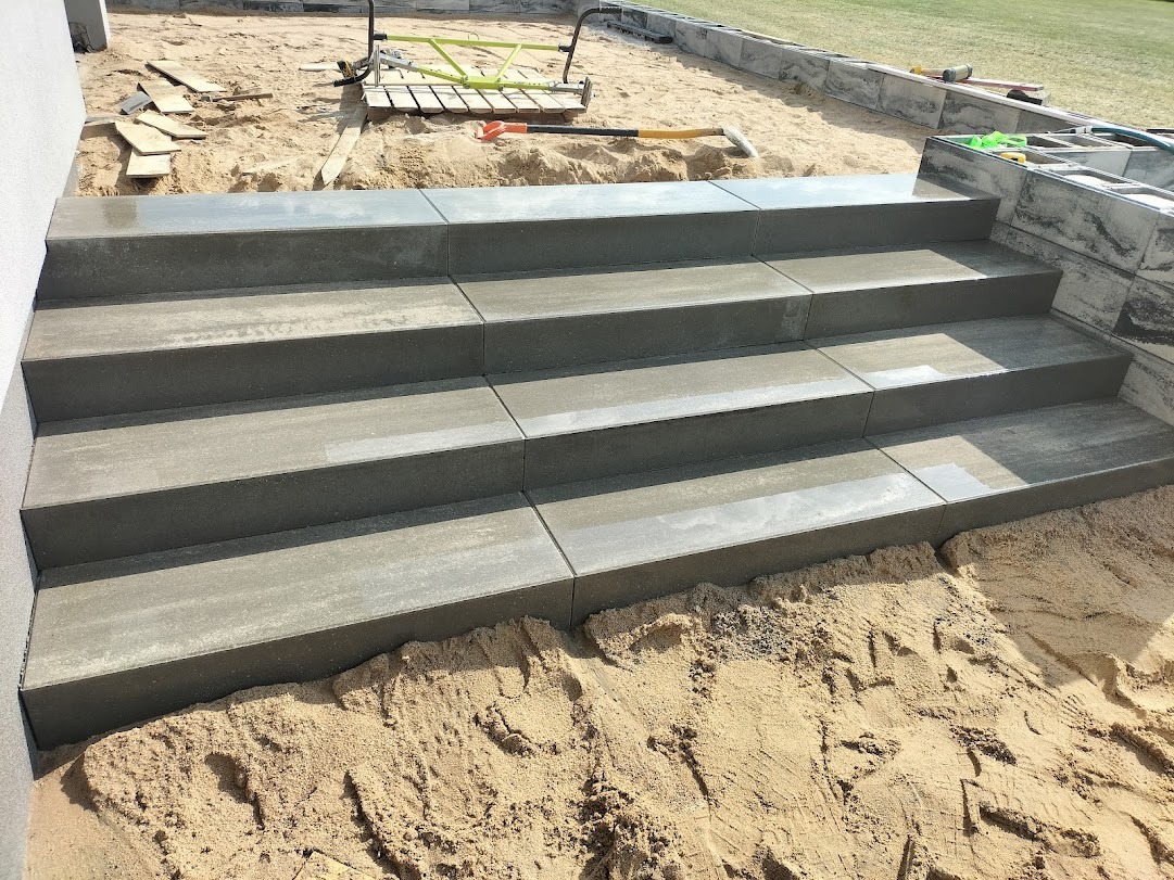 Czterostopniowe schody zewnętrzne z szarej kostki brukowej, w trakcie budowy, widoczny piasek i narzędzia.