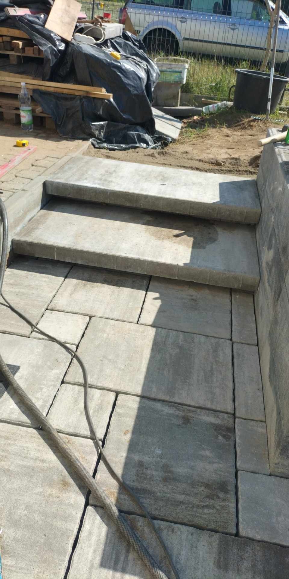 Schody i nawierzchnia z szarej kostki brukowej w trakcie układania, widoczny wąż ogrodowy, materiały budowlane w tle.
