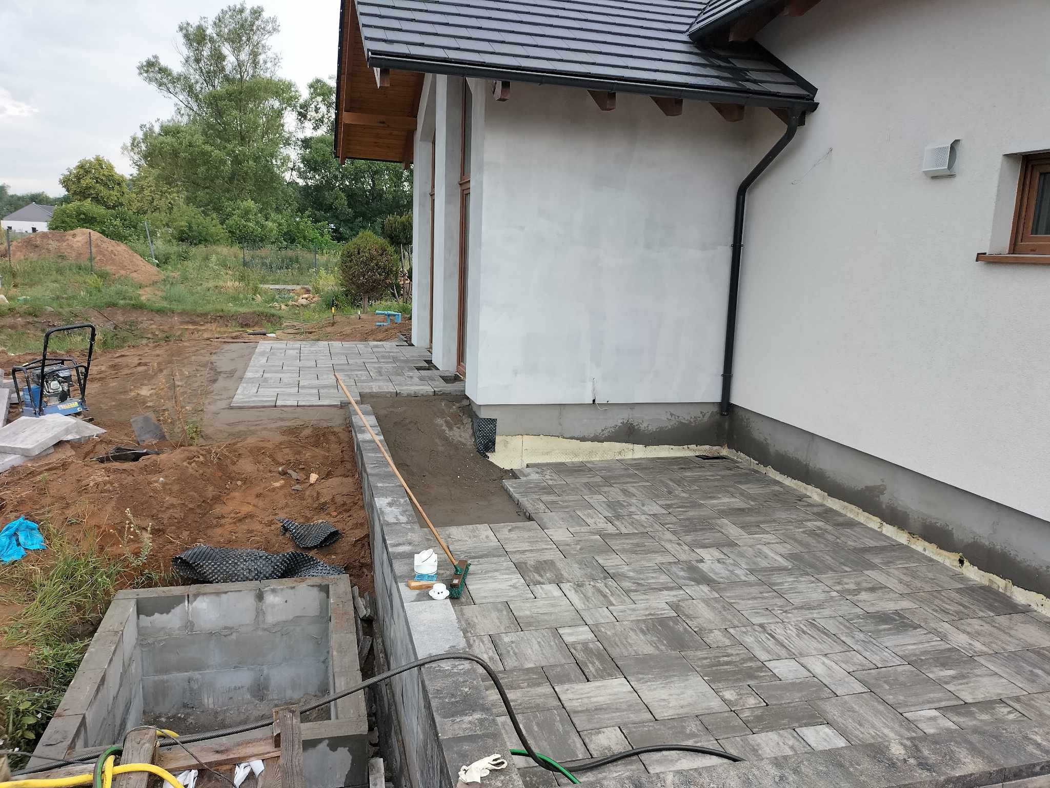 Fragment tarasu z szarej kostki, widoczny etap prac z przygotowaniem podłoża i częściowo ułożonymi płytami, obok ściana budynku z widocznym oknem i rynną oraz elementy konstrukcji drenażowej.