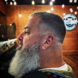 Profil mężczyzny z siwą brodą i modnym cieniowaniem włosów, widoczny na tle ceglanej ściany i oświetlenia w stylu retro w salonie.