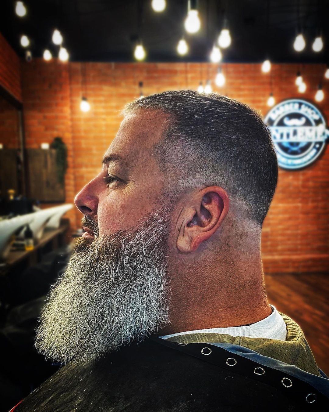 Profil mężczyzny z siwą brodą i modnym cieniowaniem włosów, widoczny na tle ceglanej ściany i oświetlenia w stylu retro w salonie.