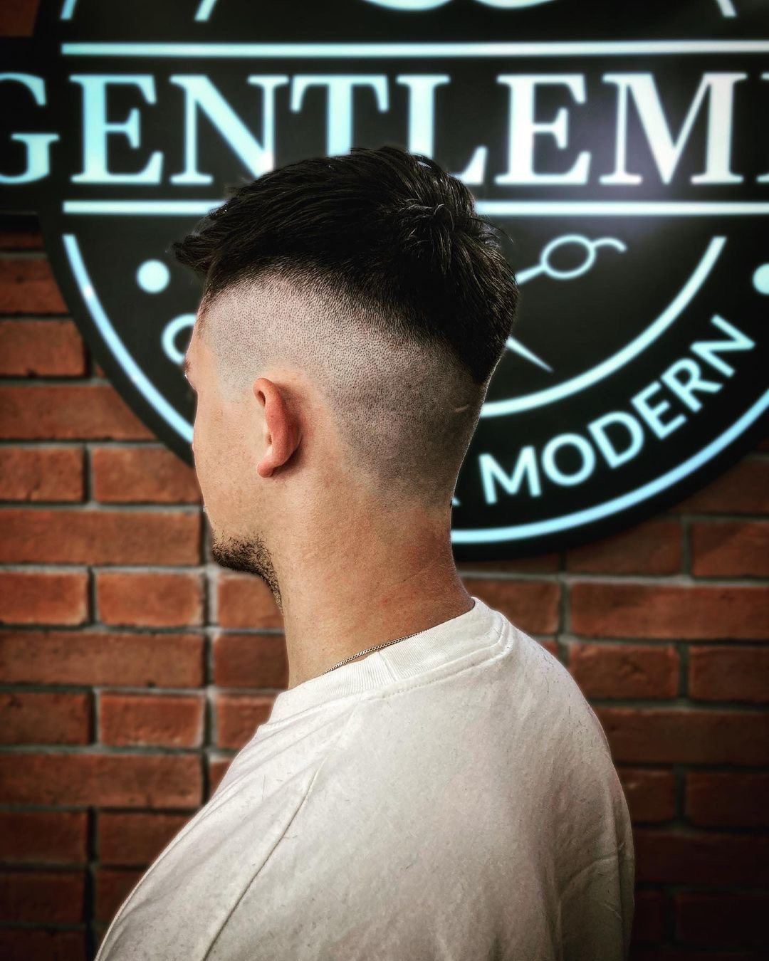 Męskie cięcie włosów z wycieniowanymi bokami, widok z profilu na tle ściany z cegły i podświetlanego logo salonu.