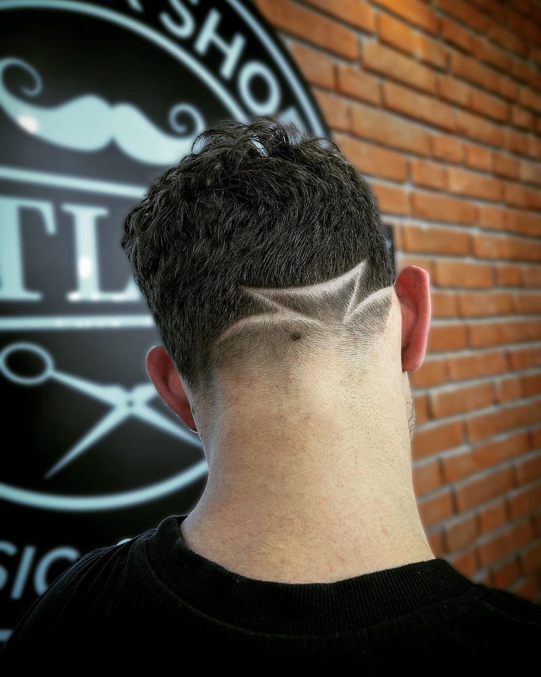 Wykonywanie wzoru geometrycznego na krótko ostrzyżonych włosach na karku mężczyzny, widok z tyłu, w tle ściana z cegły i logo salonu.