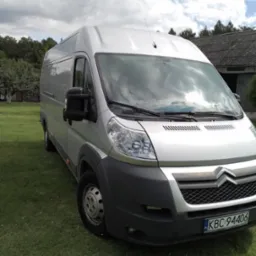 Srebrny bus marki Citroen, zaparkowany na trawniku przed budynkiem gospodarczym z szarą dachówką, widoczne odbicie chmur na przedniej szybie.
