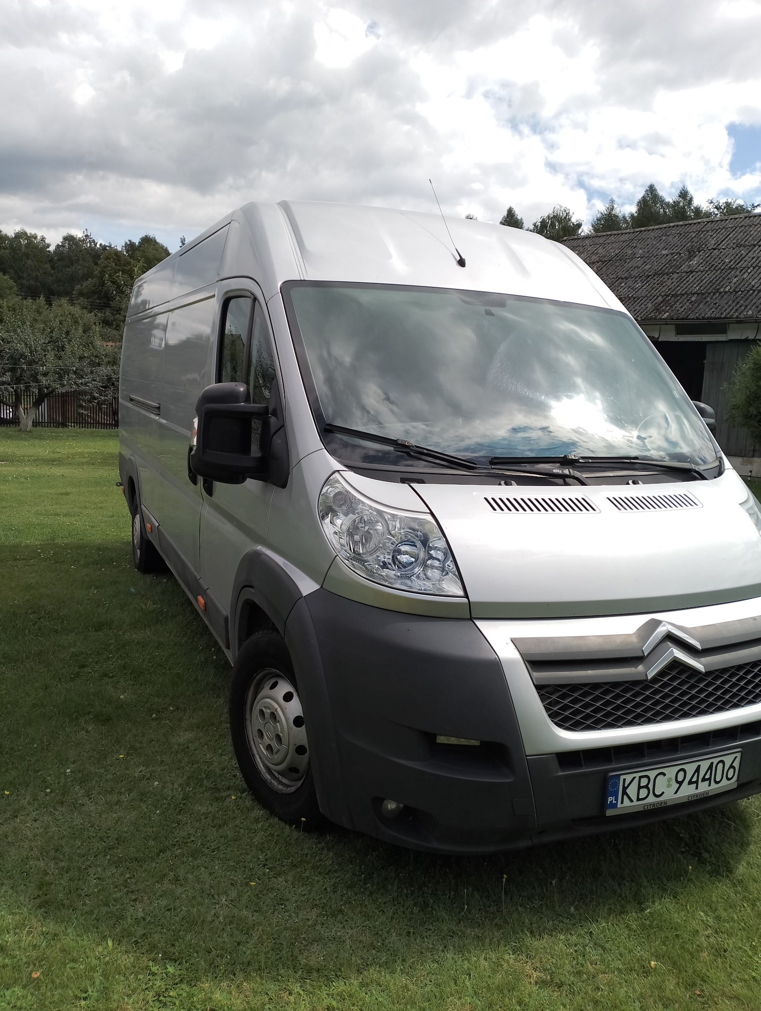 Srebrny bus marki Citroen, zaparkowany na trawniku przed budynkiem gospodarczym z szarą dachówką, widoczne odbicie chmur na przedniej szybie.