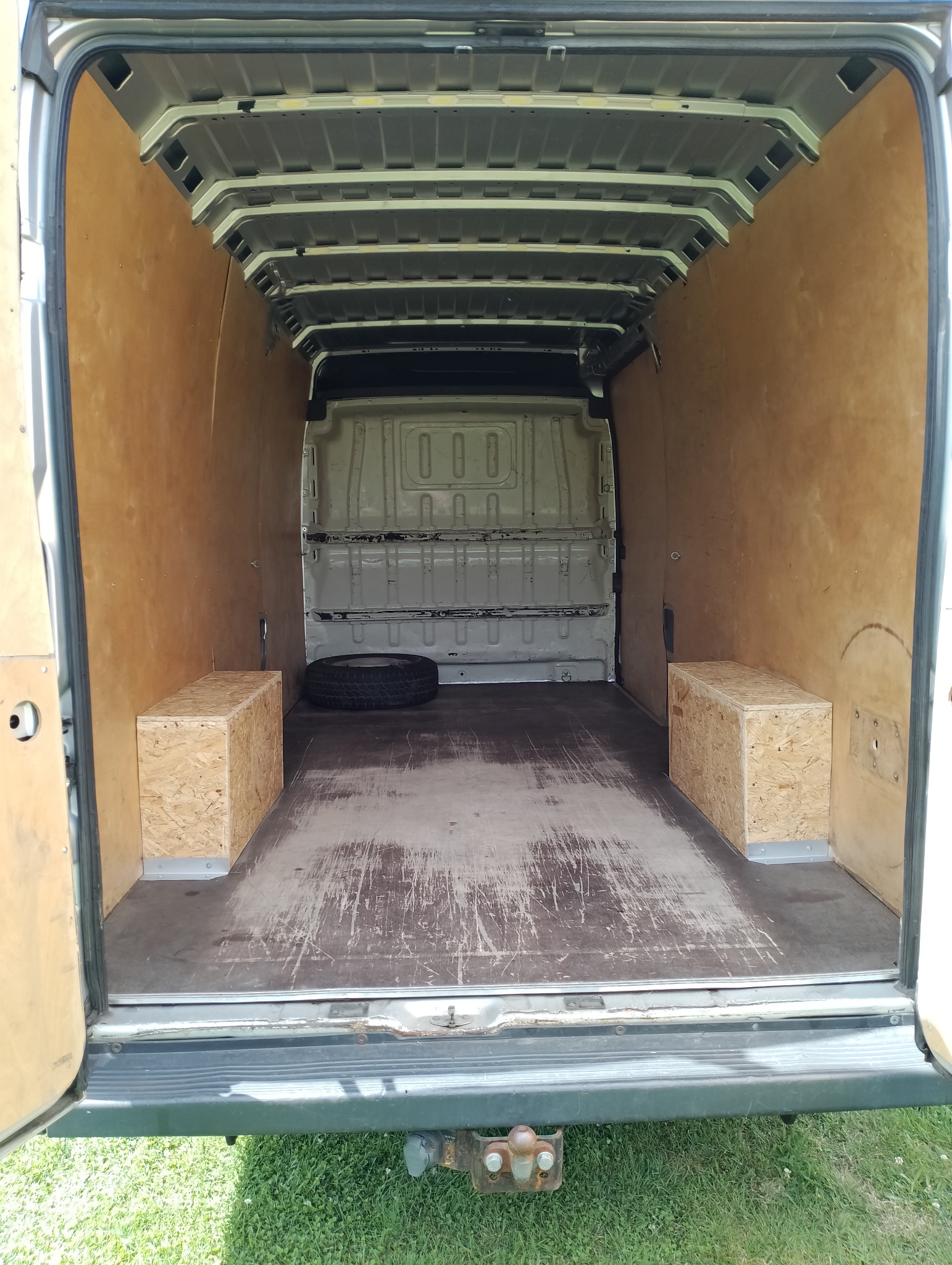 Wnętrze dostawczego samochodu wyłożone sklejką, z dwoma skrzyniami z płyty OSB po bokach i oponą w tle.