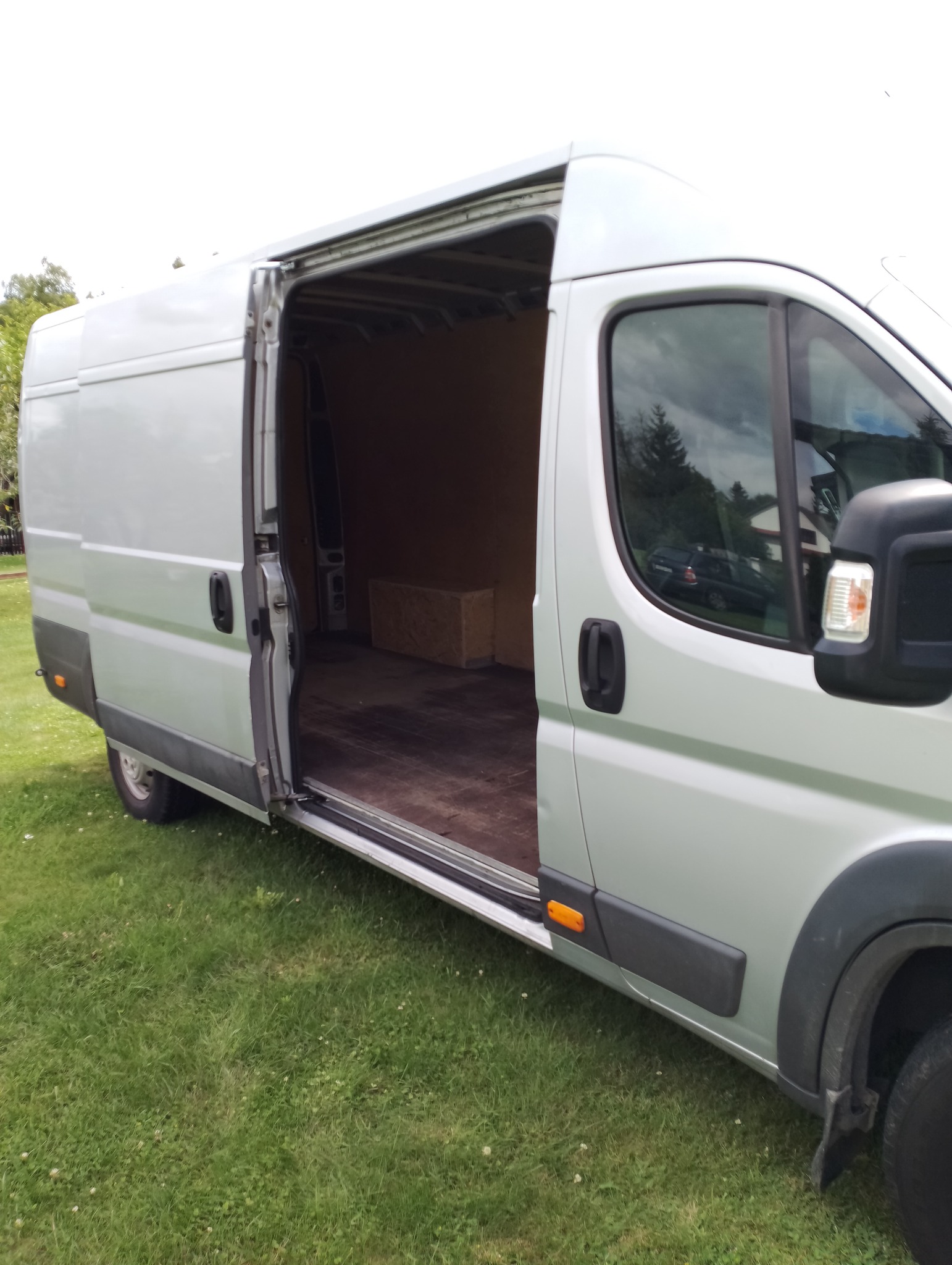 Srebrny bus z otwartymi drzwiami bocznymi, wnętrze puste z widoczną skrzynią, zaparkowany na trawniku.