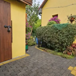 Urokliwy zakątek ogrodu z kostką brukową, drewnianą bramą i starannie przystrzyżonym żywopłotem ozdobionym koronkową serwetą, żółta elewacja budynku w tle.