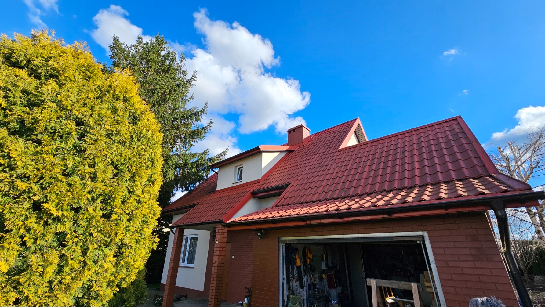 Dom jednorodzinny z czerwoną dachówką, widok z dołu, z otwartym garażem i bujnym, żółtym tujem po lewej stronie, na tle błękitnego nieba z chmurami.