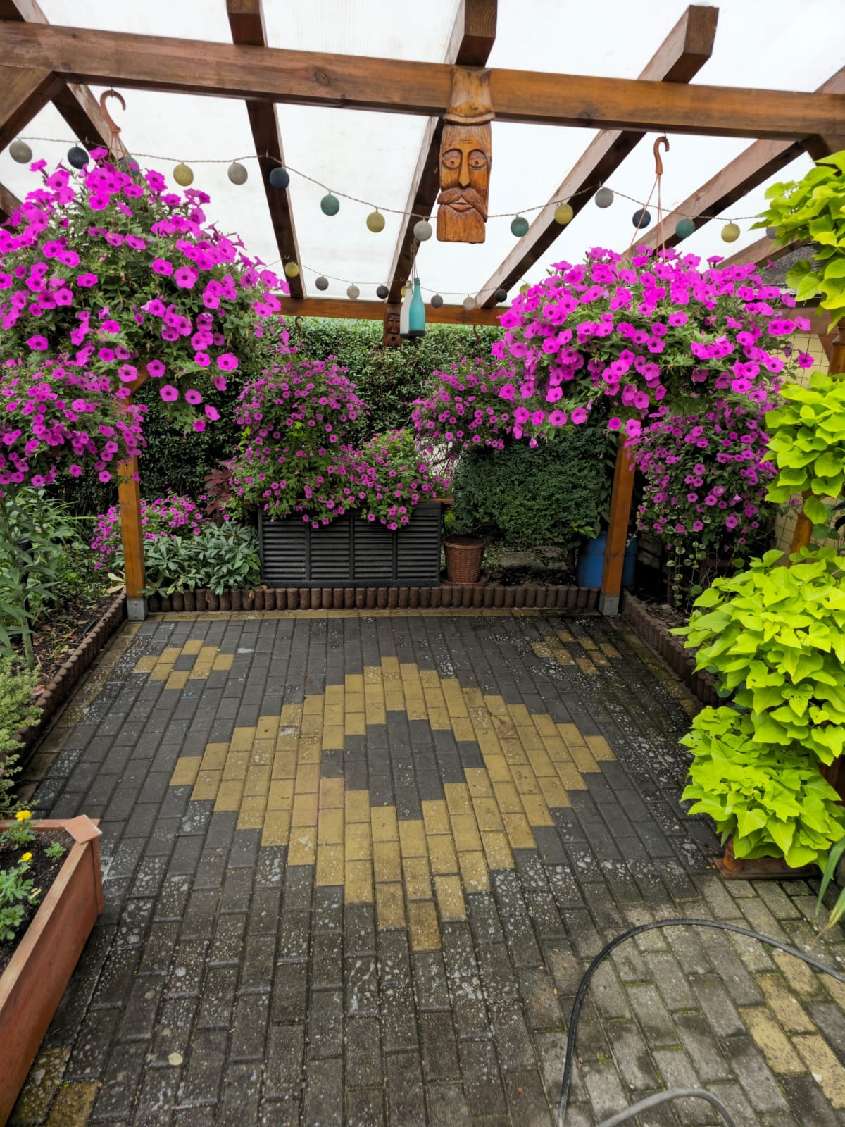 Ogród z kostki brukowej z wzorem w romby, otoczony wiszącymi koszami z różowymi kwiatami, drewnianą pergolą z dekoracją w postaci rzeźby twarzy i sznurem lampek.