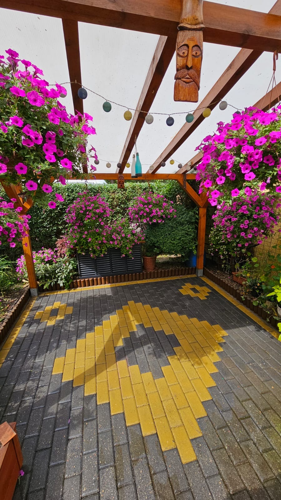 Ogród z altaną, wiszącymi kwiatami i kostką brukową ułożoną we wzór geometryczny, ozdobiona drewnianą rzeźbą i girlandą świetlną.