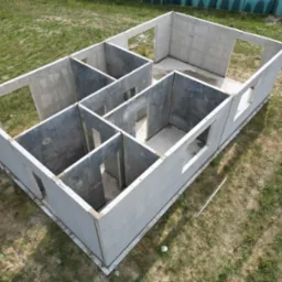 Surowe ściany parterowego domu z keramzytu na fundamencie, widok z góry na trawnik z zieloną trawą i śladami opon, zarys przyszłych okien i drzwi.