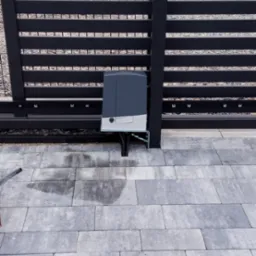 Szara, pozioma brama przesuwna z automatyką zamontowaną na słupku, widoczna kostka brukowa i fragment niebieskiej pianki izolacyjnej.