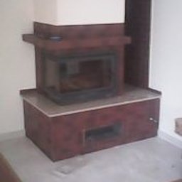 KOMPLEX MB - Wykończony kominek z brązowymi kaflami i szarym blatem w rogu pokoju. Widoczny fragment podłogi wyłożonej szarymi płytami. Całość utrzymana w ciepłej tonacji.