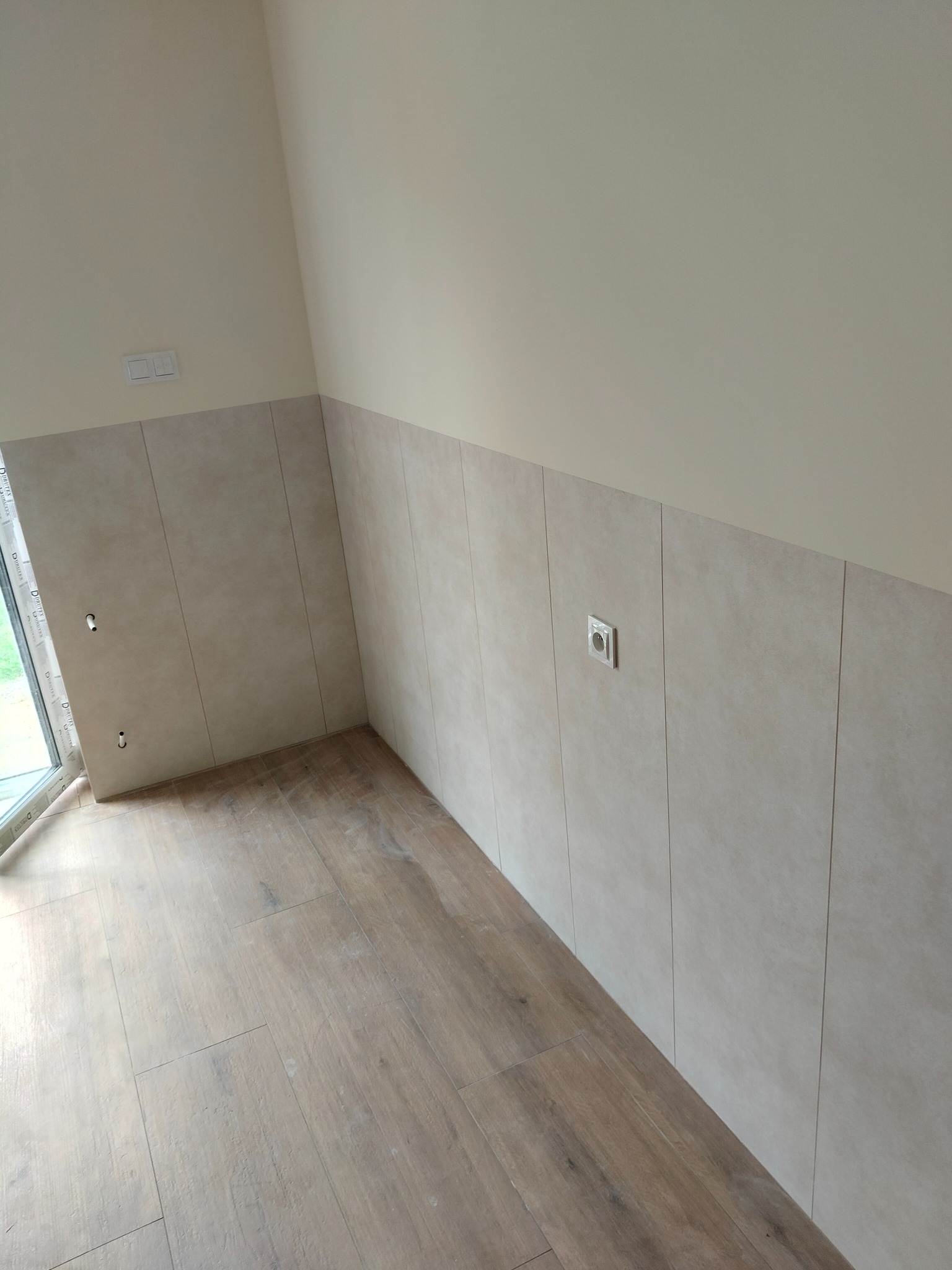 Wykończone wnętrze: kremowe płytki ścienne do połowy wysokości, drewnopodobna podłoga, białe gniazdko elektryczne, okno. Neutralna paleta barw, minimalistyczny design.