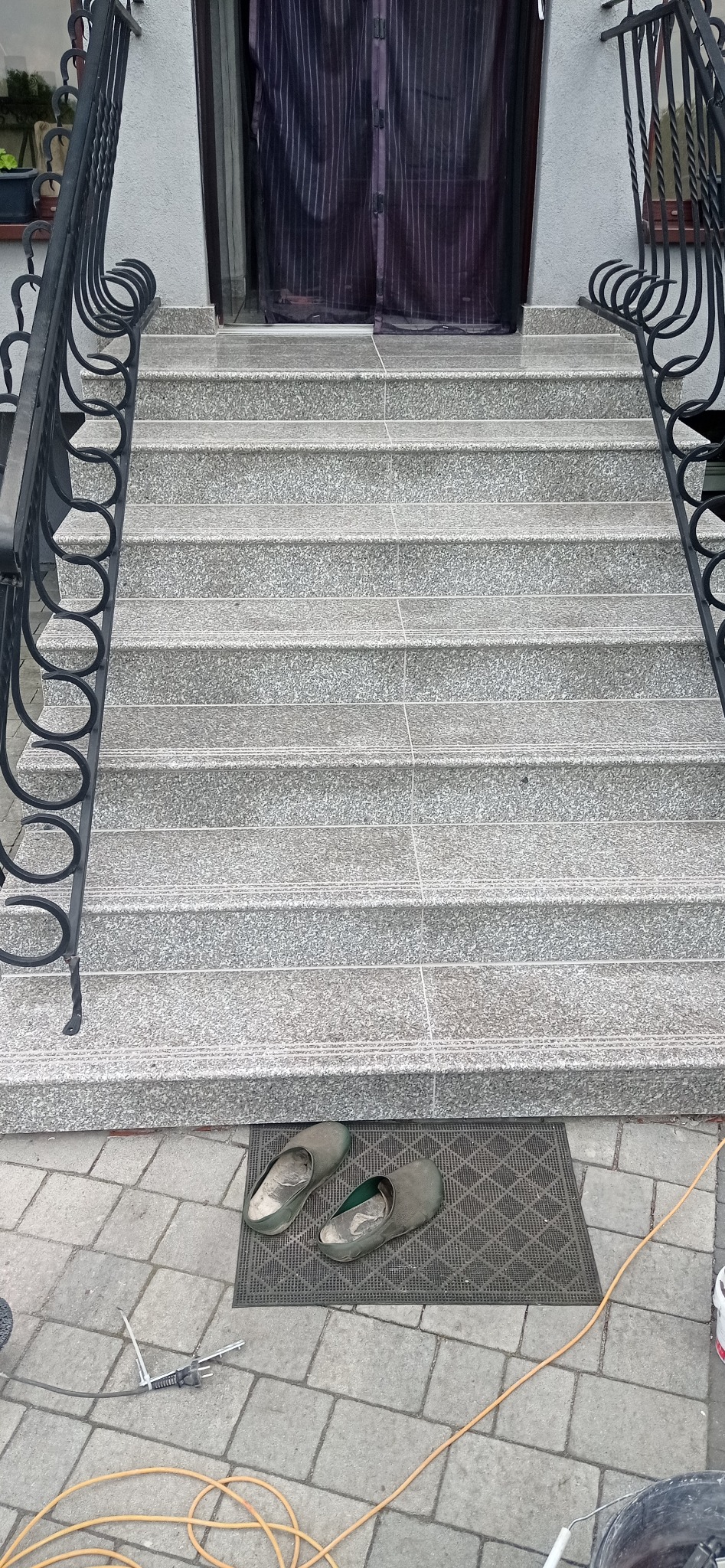 Granitowe schody wejściowe z czarną, kutą balustradą. Na wycieraczce leżą robocze klapki, obok narzędzia i kabel. Widok z góry na wykończone schody.