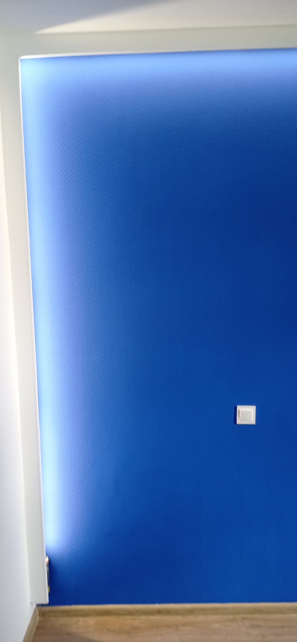 Niebieska ściana z delikatnym wzorem w jodełkę, oświetlona od góry. Widoczny fragment listwy przypodłogowej i włącznik światła.