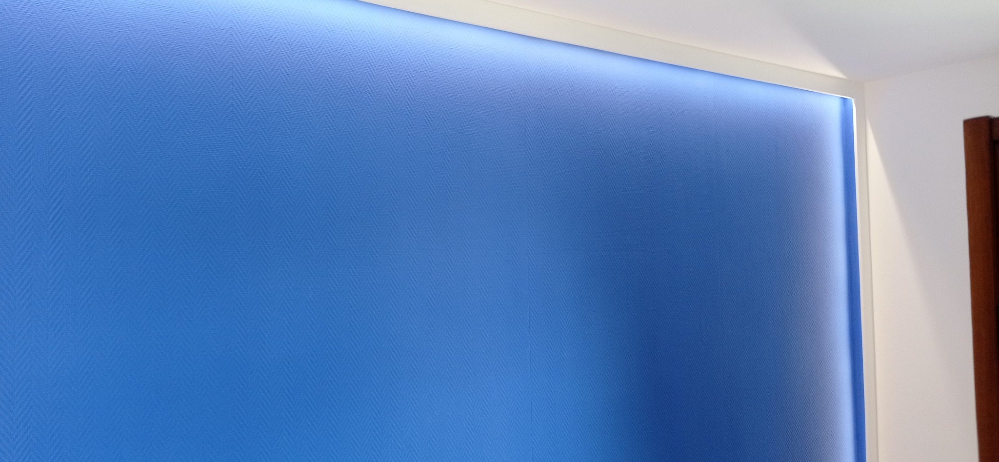 Niebieska ściana z delikatną teksturą w jodełkę, oświetlona od góry listwą LED, widoczny fragment białej ściany i drzwi.