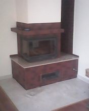 Wykończony kominek z brązowymi kaflami i szarym blatem w rogu pokoju. Widoczny fragment podłogi wyłożonej szarymi płytami. Całość utrzymana w ciepłej tonacji.