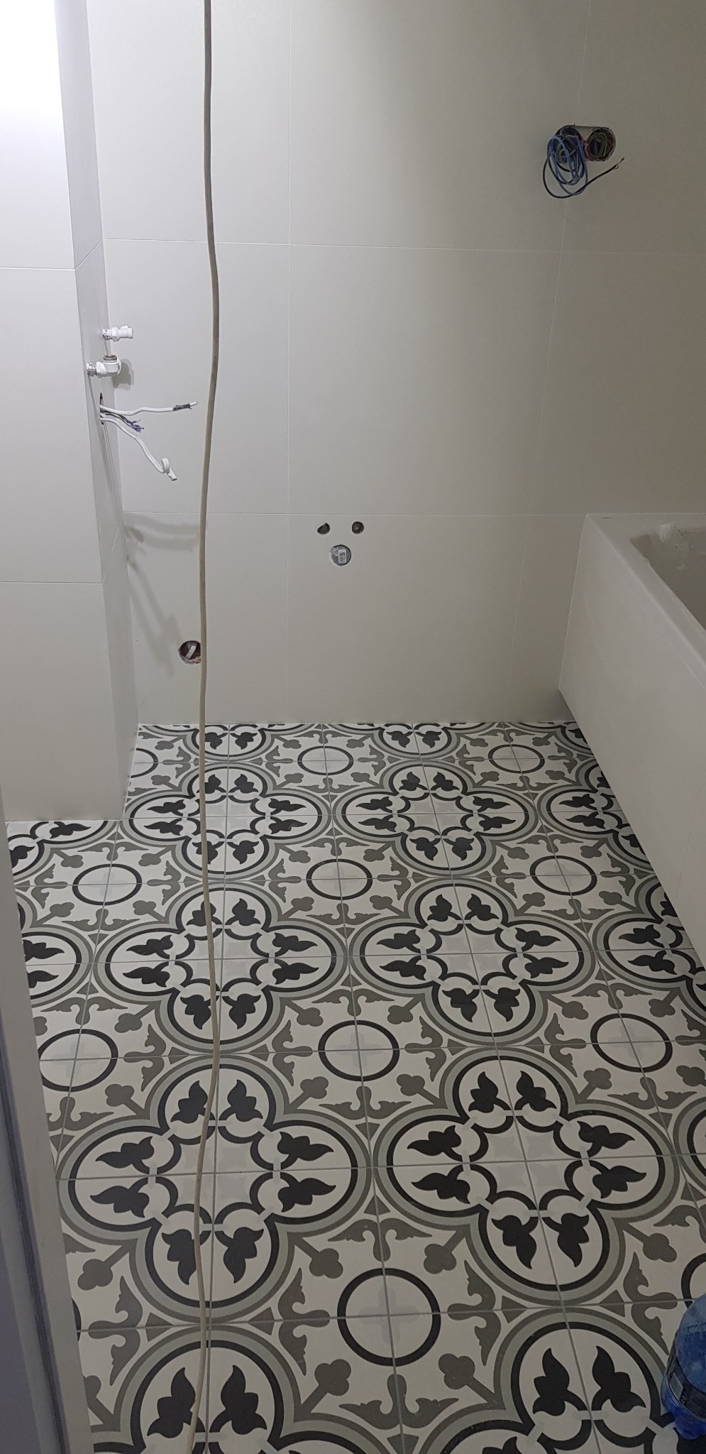 Łazienka w trakcie remontu: białe płytki ścienne, wzorzysta podłoga, widoczne wystające kable i rury.