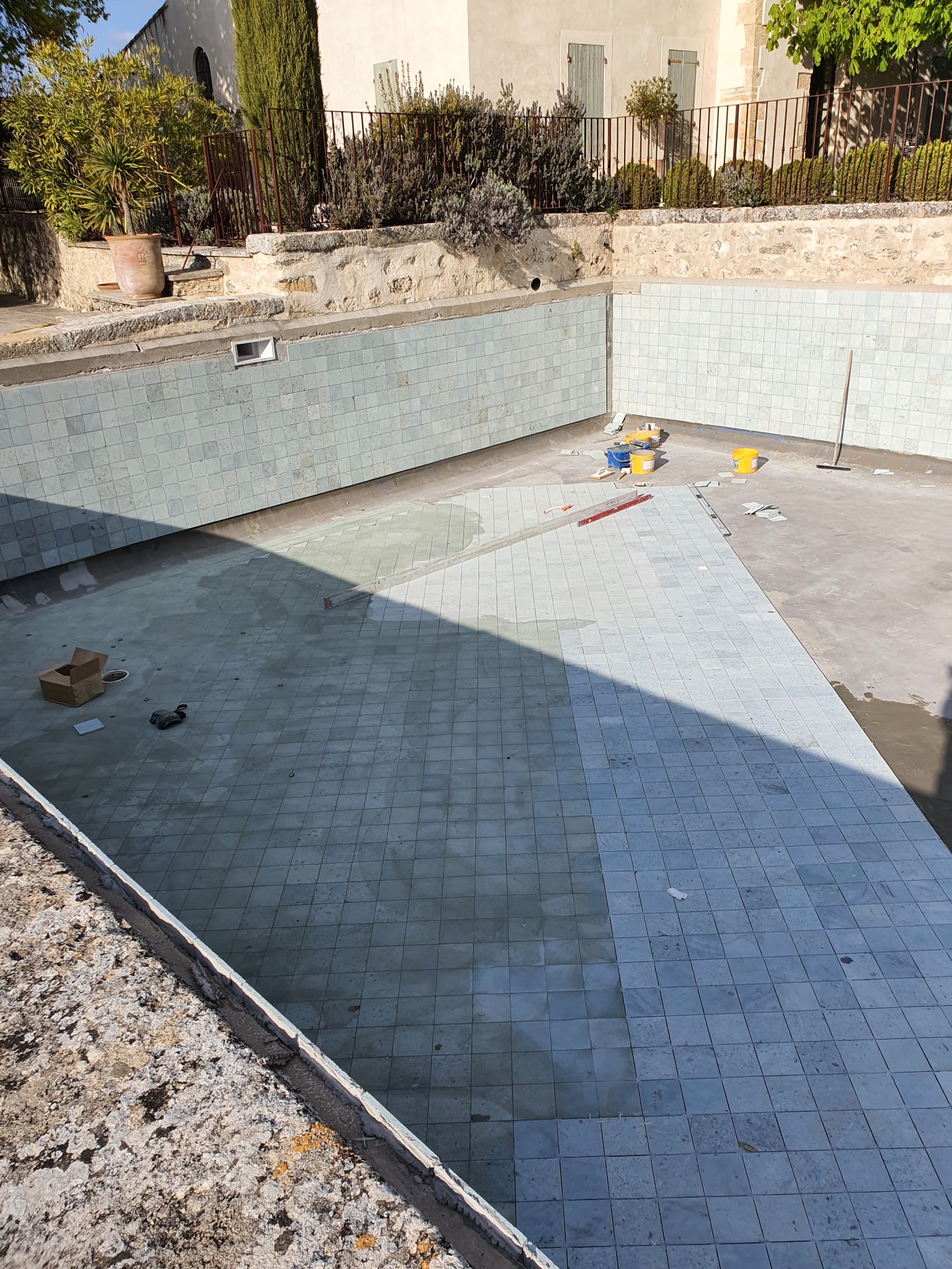 Basen w trakcie renowacji, z częściowo ułożonymi, jasnoniebieskimi płytkami; widoczne narzędzia i wiaderka z zaprawą.