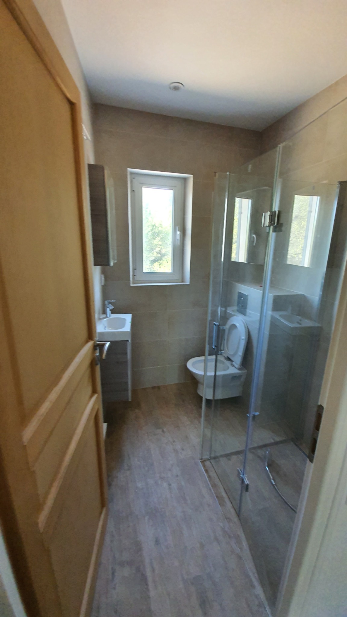 Nowoczesna łazienka z kabiną prysznicową ze szkła, umywalką z szafką, toaletą i oknem z widokiem na zieleń, wykończona płytkami w odcieniach beżu i drewnopodobną podłogą.