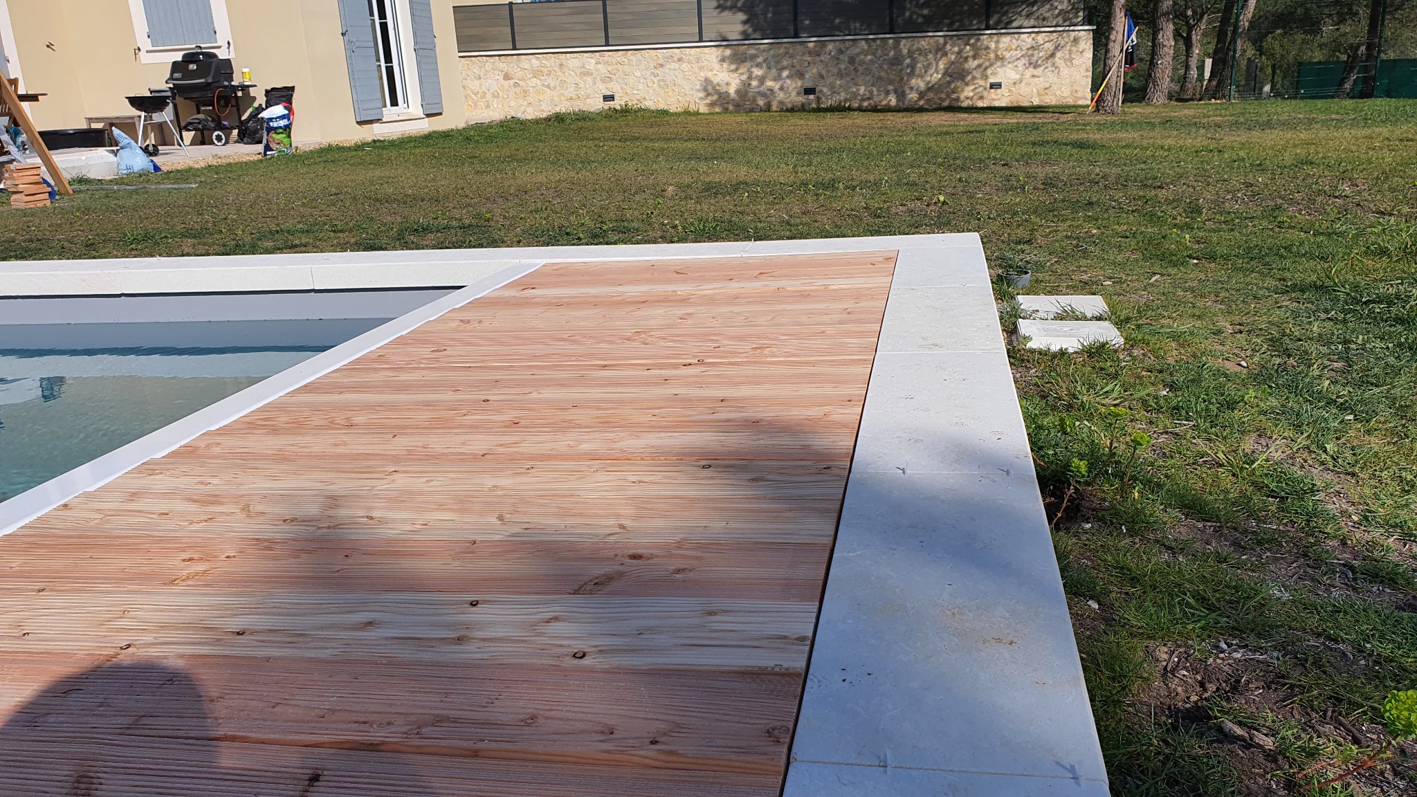 Drewniany taras przy basenie z jasnymi płytami obrzeżowymi i trawnikiem w tle, widok z góry.