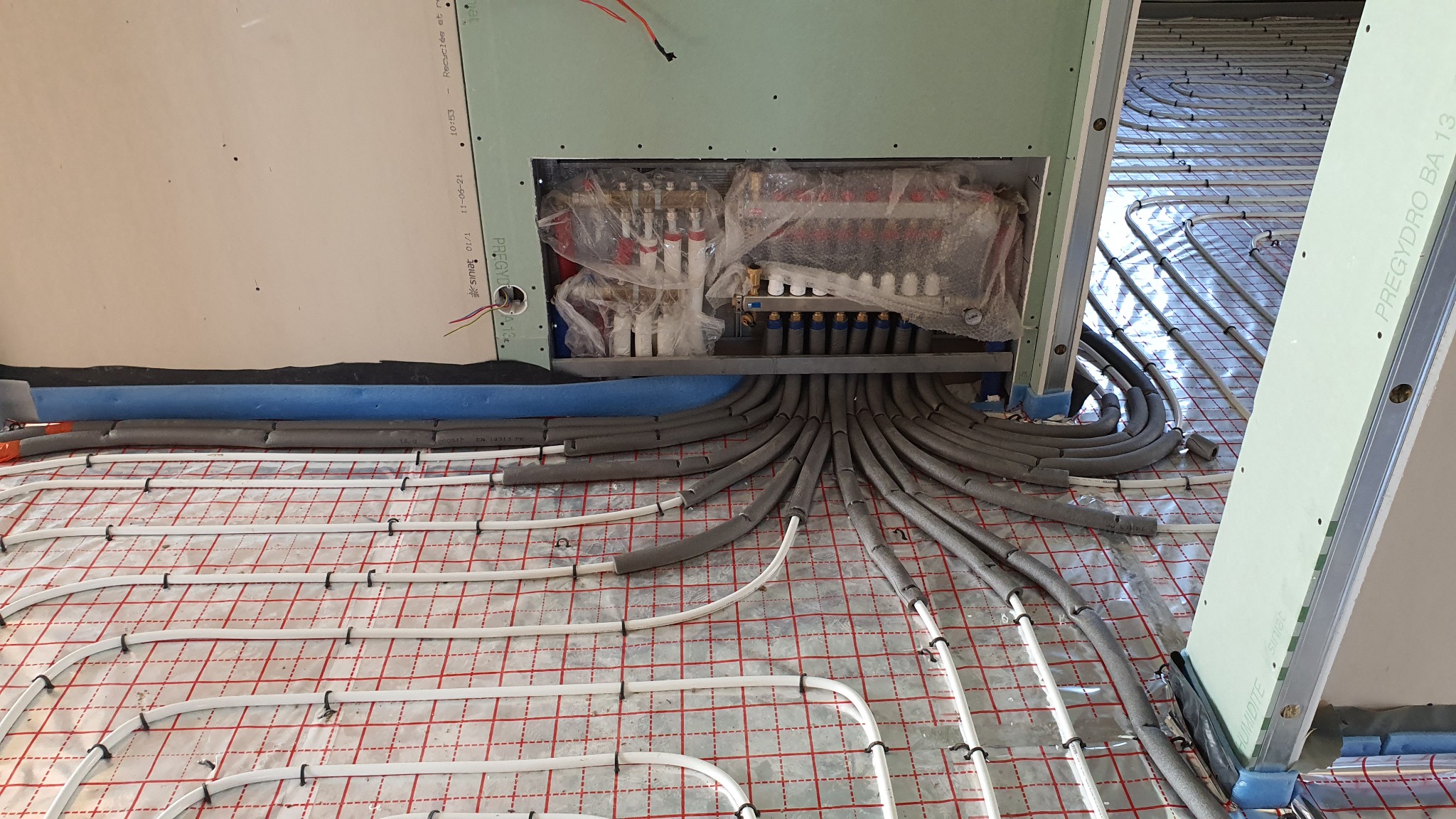 Rozdzielacz ogrzewania podłogowego, rury grzewcze ułożone na folii z siatką, izolowane rury doprowadzające, ściany w trakcie montażu płyt kartonowo-gipsowych.